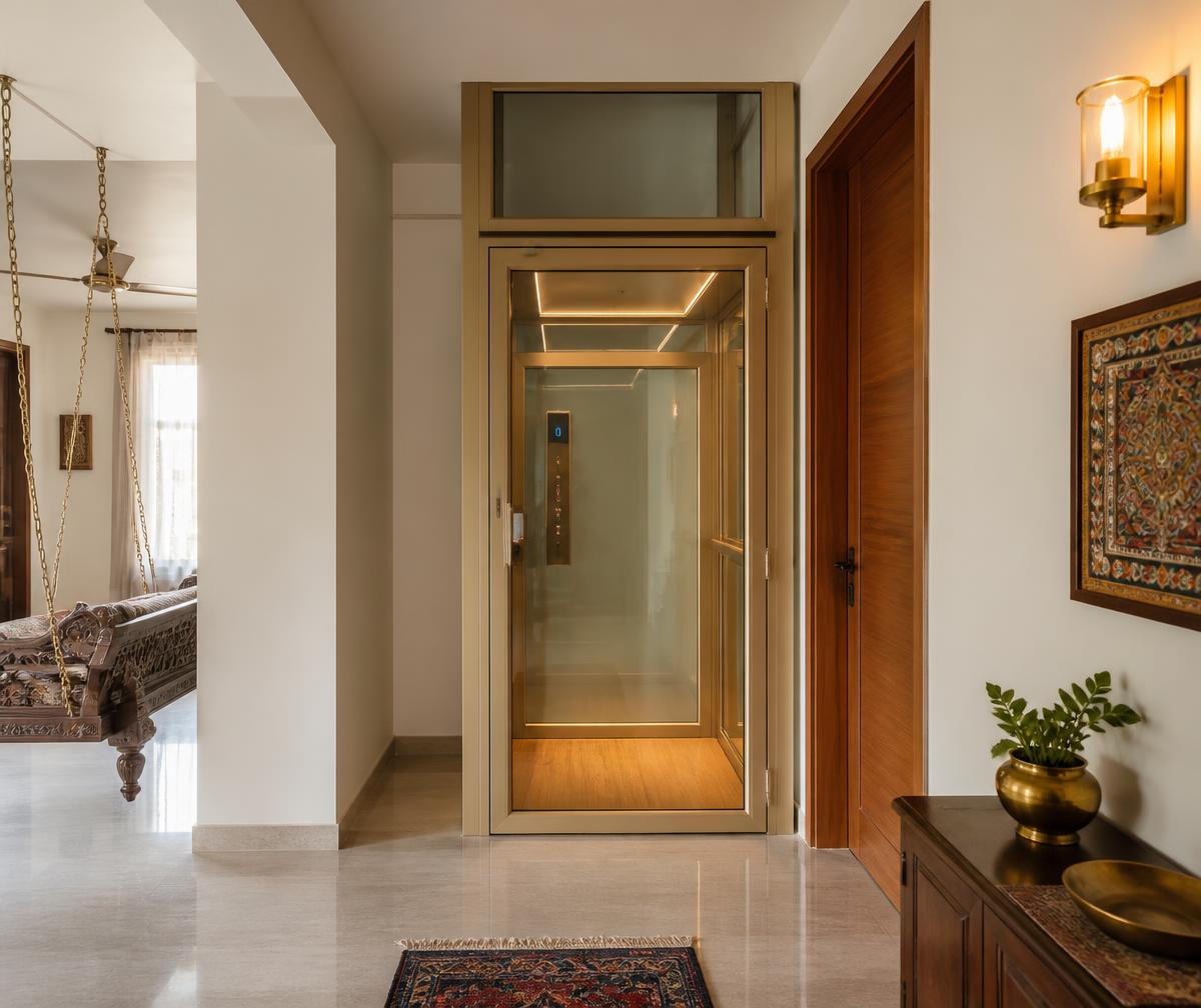 Glass and aluminium home lift at the end of a hallway beside a teak door in an Indian family home